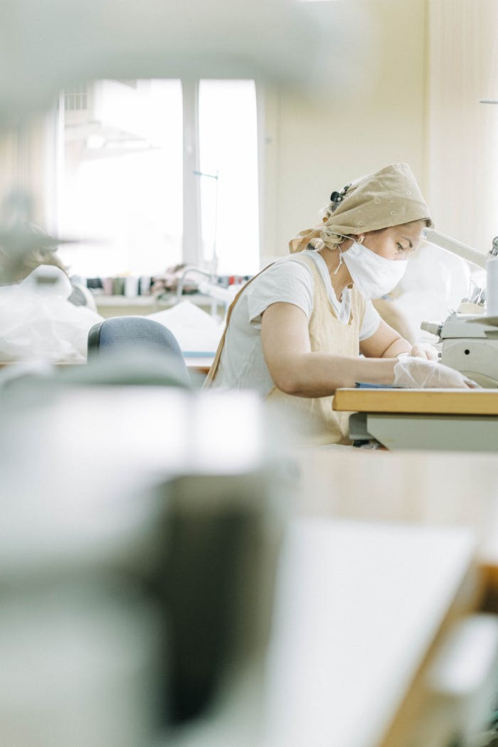 Female worker wearing face mask and apron, sewing in a factory environment.